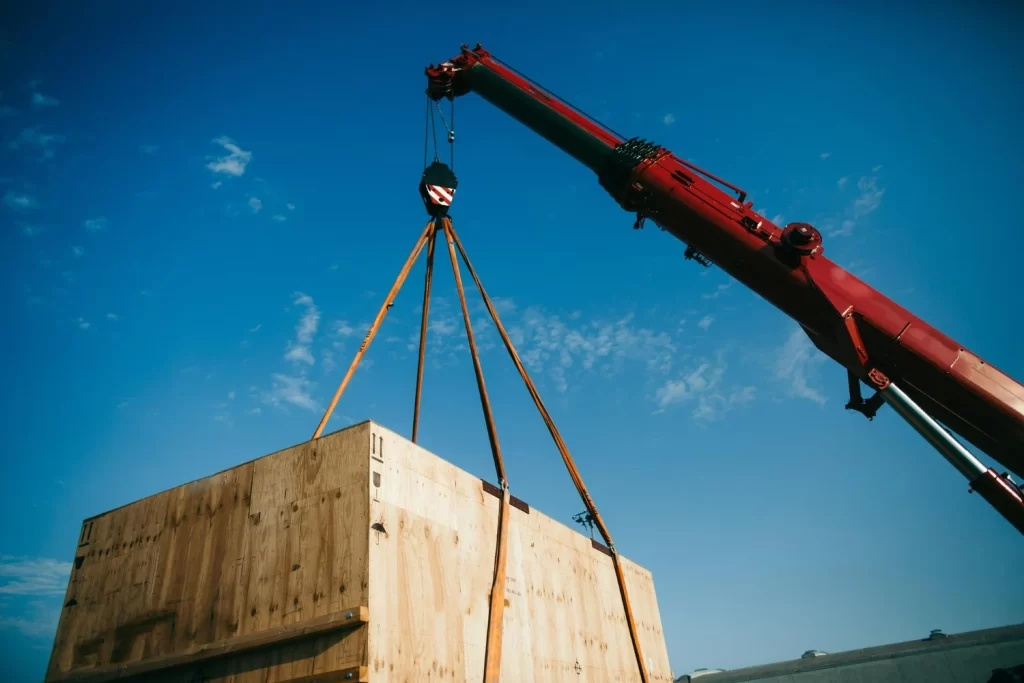 Guindaste equipado com cintas de amarração faz o içamento de carga pesada, movimentando um grande container de madeira.