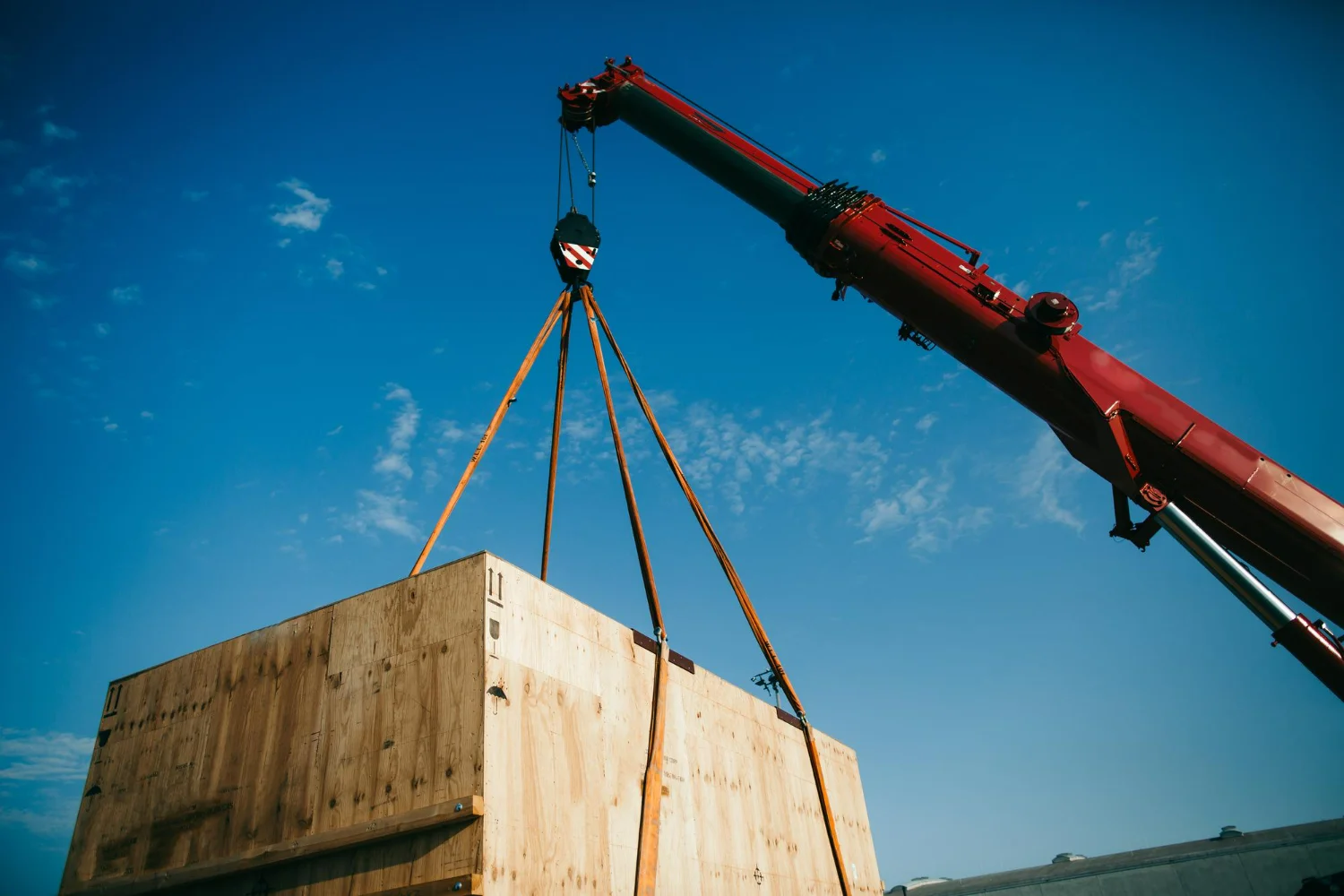 Guindaste equipado com cintas de amarração faz o içamento de carga pesada, movimentando um grande container de madeira.