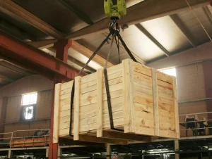 Caixa de madeira sendo içada com cintas em armazém, destacando o fator de segurança na elevação de cargas